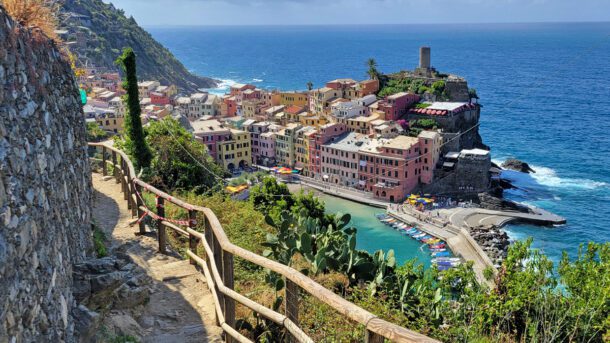 The Cinque Terre, or “Five Lands,” is one of Italy’s most breathtaking destinations. These five centuries-old fishing villages—Monterosso al Mare, Vernazza, Corniglia, Manarola, and Riomaggiore—cling to the cliffs of the Ligurian coast. These gorgeous villages of Cinque Terre are connected by hiking trails, trains, and boats. Cars are not allowed within the villages, preserving their authenticity and charm. Visitors can hop on the train that winds through tunnels linking each town, sail along the glittering Ligurian Sea for coastal views, or trek the famous Sentiero Azzurro (Blue Trail) for unforgettable panoramas. Each village has its own history, atmosphere, and attractions, creating a once-in-a-lifetime journey through color, culture, and cuisine. Here are the five gorgeous villages of Cinque Terre, listed from north to south. Monterosso al Mare The largest of the Cinque Terre villages, Monterosso al Mare dates back to the 11th century and was once heavily fortified to defend against pirate attacks. Today, it blends history with seaside leisure. It’s the only village with a wide sandy beach, making it perfect for swimming and sunbathing. Divided into the medieval Old Town and the modern New Town, Monterosso offers both historic charm and resort-style amenities. The Old Town features narrow alleys, the Gothic Church of San Giovanni Battista, and the Capuchin Monastery with sweeping sea views. In the New Town, you’ll find a lively promenade filled with cafes, gelaterias, and shops. Lemon groves and vineyards surrounding the village produce some of Liguria’s best flavors, from wine to limoncello. Whether you’re relaxing by the sea, hiking coastal trails, or exploring its historic heart, Monterosso is a must-see. Things to See and Do in Monterosso al Mare: • Relax on the sandy beach—the largest in Cinque Terre • Visit the Church of San Giovanni Battista with its black-and-white striped façade • Climb to the Capuchin Monastery for views of the village and coast • Stroll along the seafront promenade lined with shops and cafes • See the Giant Statue of Neptune, a dramatic cliffside sculpture • Hike the coastal trail to Vernazza for unforgettable panoramas Vernazza Founded around 1000 AD, Vernazza is considered the jewel of Cinque Terre. Once a vital port for the Republic of Genoa, it prospered as a fishing and trade hub. Its natural harbor, framed by colorful houses, remains the heart of the village. Vernazza is crowned by the 15th-century Doria Castle, built to protect against pirates and now offering incredible views of the Ligurian Sea. At the center of the village is Piazza Marconi, where locals and visitors gather at outdoor cafes. The Church of Santa Margherita di Antiochia, dating back to the 14th century, stands right at the water’s edge. Vernazza also has a small sandy beach, making it perfect for a refreshing swim. The cobblestone streets are dotted with gelato shops and artisan boutiques, while the surrounding hillsides invite exploration via hiking trails. With its mix of history, culture, and natural beauty, Vernazza captures the spirit of Cinque Terre. Things to See and Do in Vernazza: • Climb to Doria Castle for panoramic views • Relax at the harborfront piazza with a glass of wine • Visit the Church of Santa Margherita di Antiochia • Swim or sunbathe at the small sandy beach • Explore artisan shops and sample local gelato • Hike the famous trail between Vernazza and Monterosso Corniglia Perched on a cliff 100 meters above the sea, Corniglia is the only Cinque Terre village not directly on the water. With roots stretching back to Roman times, Corniglia has long been a wine-producing hub, surrounded by terraced vineyards that yield Sciacchetrà, a sweet regional wine. To reach it, visitors climb the Lardarina, a staircase of more than 350 steps, or take a shuttle bus from the train station. Once at the top, the reward is sweeping views of the coastline in both directions. Corniglia is quieter and less touristy than the other villages, making it feel more authentic. The village square, Largo Taragio, is lined with cozy cafes and shops, perfect for relaxing with coffee or wine. The Church of San Pietro, built in 1334, is a striking example of Gothic-Ligurian architecture. For hikers, Corniglia is a gateway to both coastal and hillside trails. Its elevated perch, rustic charm, and peaceful atmosphere make it a haven for travelers seeking a slower pace. Things to See and Do in Corniglia: • Climb the Lardarina staircase or ride the shuttle up from the train station • Visit the Church of San Pietro with its Gothic design • Relax in Largo Taragio, the main village square • Explore nearby vineyards and sample Sciacchetrà wine • Enjoy panoramic views of the coast from multiple viewpoints • Hike trails leading to Vernazza or Manarola Manarola Manarola, founded in the 12th century, is perhaps the most photographed of the Cinque Terre villages. Its colorful houses cascade dramatically down the cliffs toward a small harbor, creating one of Italy’s most iconic views. Historically, Manarola has been a center for viticulture, and the terraced hillsides above still produce the prized Sciacchetrà dessert wine. The village is filled with narrow lanes, lively cafes, and charming shops. For romance, take the Via dell’Amore, the “Path of Love,” a scenic walkway that once linked Manarola to Riomaggiore along the coast. Adventurers will love cliff jumping or swimming in the harbor, while hikers can ascend trails into the hills for stunning panoramas of the town and sea. At sunset, Manarola glows with golden light, reflecting off the houses and water in a dazzling display. Whether sipping wine on a terrace, photographing its iconic skyline, or exploring its vineyards, Manarola offers an unforgettable mix of beauty and tradition. Things to See and Do in Manarola: • Stroll the scenic Via dell’Amore (when open) • Swim or cliff jump in the small harbor • Photograph the iconic cliffside houses at sunset • Visit the Church of San Lorenzo, built in 1338 • Sample local Sciacchetrà wine from nearby vineyards • Hike into the hills for sweeping views Riomaggiore Riomaggiore, the southernmost Cinque Terre village, dates back to the 13th century. It was founded by settlers escaping the hills, who built terraced vineyards and tall, colorful houses stacked dramatically down toward the harbor. Its main street, Via Colombo, buzzes with shops, cafes, and trattorias serving local specialties. At the top of the village, the Church of San Giovanni Battista (1340) displays Gothic architecture and stunning views, while the Castle of Riomaggiore offers a glimpse into its defensive past. Down below, the marina is perfect for watching fishermen or renting kayaks to explore hidden coves. At sunset, Riomaggiore becomes magical, as travelers and locals gather on the rocks by the sea with wine to watch the horizon glow in shades of orange and purple. With its vibrant streets, historic landmarks, and lively atmosphere, Riomaggiore embodies the soul of the Cinque Terre. Things to See and Do in Riomaggiore: • Wander Via Colombo, the main street lined with shops and cafes • Visit the Church of San Giovanni Battista (1340) • Explore the Castle of Riomaggiore and its history • Relax at the marina or rent a kayak for coastal exploration • Enjoy a glass of wine while watching the sunset from the rocks • Hike the hillside trail to Manarola What to Eat in Cinque Terre Food is central to the Cinque Terre experience. Anchovies, caught fresh daily, are served fried, marinated, or stuffed. Pesto, born in nearby Genoa, is best enjoyed over trofie pasta or spread on warm focaccia. Focaccia itself is a staple, topped with olives, onions, or cheese. Seafood risotto, grilled octopus, and farinata (a chickpea pancake) are other local favorites. Pair your meal with crisp Cinque Terre DOC white wine or indulge in Sciacchetrà, the region’s signature sweet wine. Bonus Porto Venere Porto Venere, perched on the Ligurian coast, is a hidden gem often overshadowed by nearby Cinque Terre—but its beauty is unforgettable. The town’s pastel-hued houses glow in the sunlight, perfectly reflecting in the turquoise waters of the harbor. At the edge of a dramatic cliff, the black-and-white striped Church of St. Peter rises above crashing waves, a breathtaking sight especially at sunset. Behind the waterfront, narrow medieval streets climb toward the ancient Doria Castle, offering sweeping views of the Gulf of Poets. What makes Portovenere truly special is its mix of natural beauty, colorful charm, and rich history—all wrapped in a more peaceful, authentic atmosphere than its famous neighbors. Things to do in Porto Venere: • Wander the pastel-painted harborfront and snap photos. • Visit the striking Church of St. Peter on the cliff. • Explore Doria Castle and its panoramic views. • Take a boat to Palmaria Island for hiking and swimming. • Stroll the medieval caruggi (narrow lanes) lined with shops. • Try local seafood specialties like mussels and stuffed anchovies. La Spezia Nestled along the Ligurian coast, La Spezia is a vibrant port city and the perfect gateway to the breathtaking Cinque Terre. With its colorful harbor, palm-lined promenades, and lively piazzas, La Spezia offers the charm of Italy without the heavy crowds. Stroll through the historic old town, explore fascinating museums, or savor fresh seafood at a seaside trattoria. Its central location makes it the ideal base for day trips by train or boat to Cinque Terre’s famous cliffside villages. Blending authenticity, culture, and natural beauty, La Spezia is more than a stopover—it’s a destination worth discovering. Things to Do in La Spezia • Take a boat to Cinque Terre – Hop on a ferry and enjoy breathtaking views of the colorful cliffside villages from the sea. • Visit the Naval Technical Museum – Dive into Italy’s maritime heritage with fascinating ship models, artifacts, and history. • Stroll through Parco Giardini Pubblici – Relax among fountains, palm trees, and sculptures in this waterfront park. • Taste local seafood specialties – Savor Ligurian favorites like muscoli ripieni (stuffed mussels) and fresh anchovies. • Explore the Castle of San Giorgio – A medieval fortress with sweeping views of the city and an archaeological museum inside. • Wander Via del Prione – Shop, sip coffee, and enjoy gelato along La Spezia’s charming pedestrian street. Conclusion This is where history, beauty, and tradition converge. Each of the gorgeous villages of Cinque Terre tells its own story, from Monterosso’s beaches to Corniglia’s vineyards. Whether you arrive by train, boat, or footpath, these cliffside gems will leave you spellbound. Which of the five villages will capture your heart first? Click below to PIN so you can find the five gorgeous villages of Cinque Terre again: This article may contain affiliate/compensated links. For full information, please see our disclaimer. About the Author Patti Morrow is a freelance travel writer and founder of the award-winning international blog Luggage and Lipstick. TripAdvisor called her one of the “20 Baby Boomer Travel Bloggers Having More Fun Than Millennials” and she was named one of the “Top 35 Travel Blogs” in the world. She is also the star of the upcoming TV series "Destination Takeover" which is scheduled to premiere in the next few months. Patti is the author of the book “Girls Go Solo: Tips for Women Traveling Alone,” and has over 150 bylines in 40 print and online publications, including The Huffington Post, International Living Magazine, Washington Post Sunday Travel, Travel Girl, Travel Play Live Magazine, and Ladies Home Journal. She has traveled extensively through six continents looking for fabulous destinations, exotic beaches, and adventure activities for her Baby Boomer tribe.